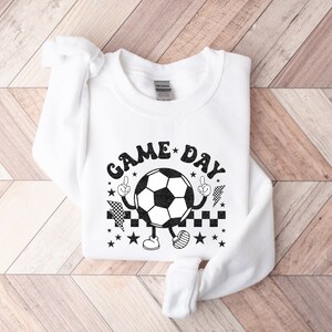 May include: White sweatshirt with a black graphic of a soccer ball with arms and legs. The soccer ball is wearing a checkered hat and has the text "GAME DAY" around it.