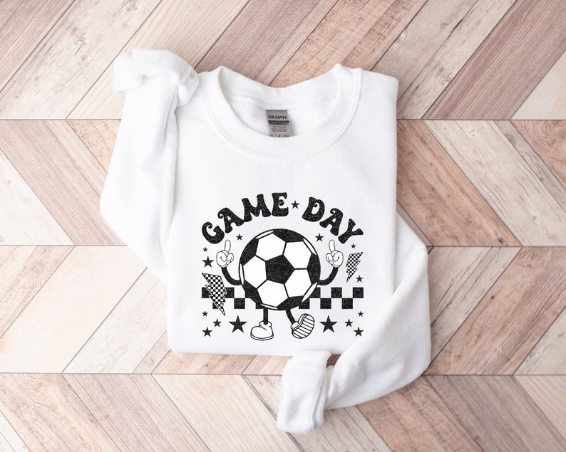 May include: White sweatshirt with a black graphic of a soccer ball with arms and legs. The soccer ball is wearing a checkered hat and has the text "GAME DAY" around it.