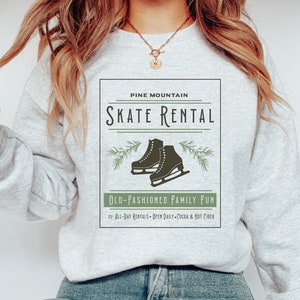 K&ouml;nnte beinhalten: Ein graues Sweatshirt mit einem grafischen Design, das ein Logo im Vintage-Stil f&uuml;r Pine Mountain Skate Rental zeigt. Das Logo enth&auml;lt ein Paar Schlittschuhe, Kiefern&auml;ste und den Text "Old-Fashioned Family Fun" mit Details zu Ganztagesmieten, t&auml;glichen &Ouml;ffnungszeiten und hei&szlig;er Schokolade und Apfelwein.