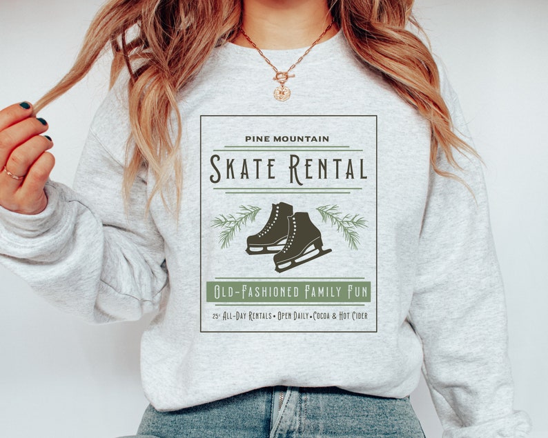 K&ouml;nnte beinhalten: Ein graues Sweatshirt mit einem grafischen Design, das ein Logo im Vintage-Stil f&uuml;r Pine Mountain Skate Rental zeigt. Das Logo enth&auml;lt ein Paar Schlittschuhe, Kiefern&auml;ste und den Text "Old-Fashioned Family Fun" mit Details zu Ganztagesmieten, t&auml;glichen &Ouml;ffnungszeiten und hei&szlig;er Schokolade und Apfelwein.