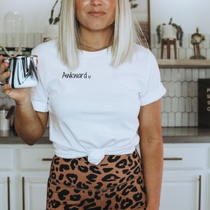 May include: A white t-shirt with the text "Awkward" and a heart symbol printed on the front. The shirt is knotted at the waist and the person wearing it is holding a black and white mug.