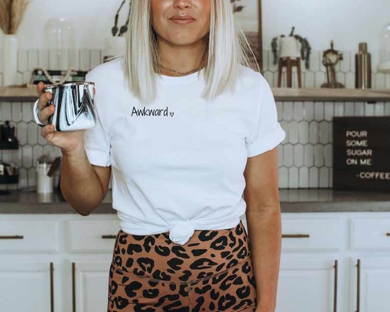 May include: A white t-shirt with the text "Awkward" and a heart symbol printed on the front. The shirt is knotted at the waist and the person wearing it is holding a black and white mug.