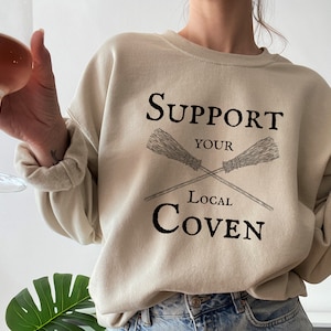 May include: A beige sweatshirt with the text "Support Your Local Coven" and a graphic of two crossed brooms.