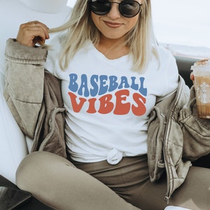 May include: A white t-shirt with a blue and red retro-style graphic that says "Baseball Vibes". The person wearing the shirt is sitting in a car and holding a coffee.