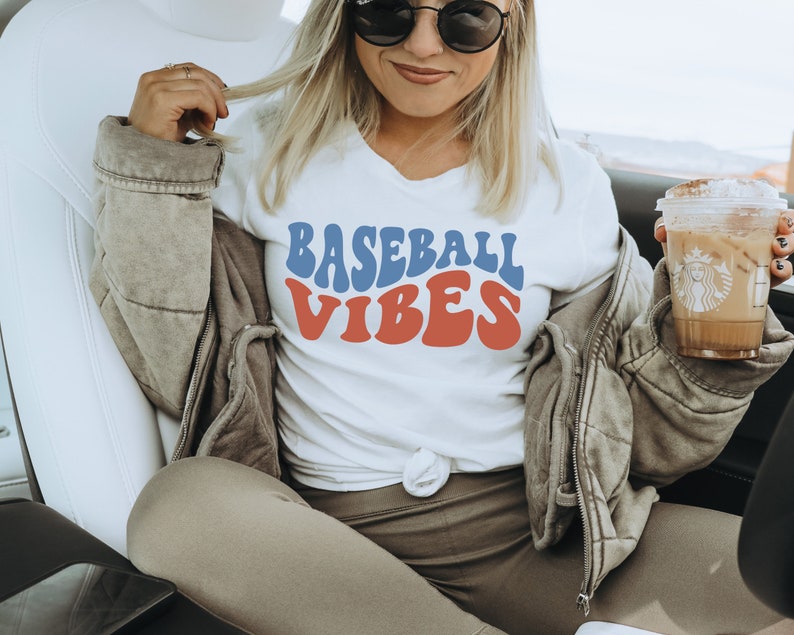 May include: A white t-shirt with a blue and red retro-style graphic that says "Baseball Vibes". The person wearing the shirt is sitting in a car and holding a coffee.
