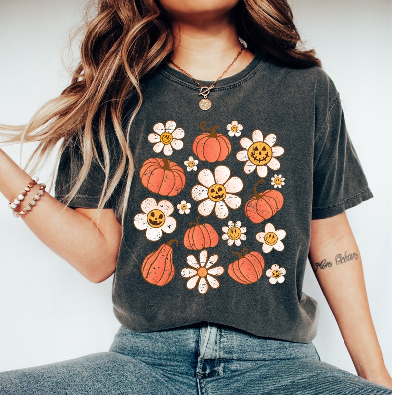 K&ouml;nnte beinhalten: Dunkelgraues T-Shirt mit einem Retro-Design mit orangefarbenen K&uuml;rbissen und G&auml;nsebl&uuml;mchen mit l&auml;chelnden Gesichtern. Das Shirt hat einen Vintage-Look, ideal f&uuml;r Herbst oder Halloween. Das Design ist mittig auf der Vorderseite des Shirts platziert.
