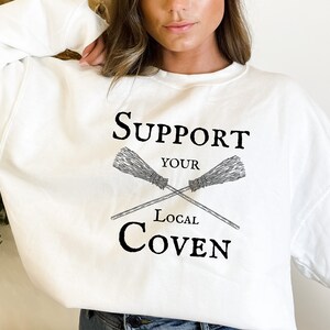 May include: White sweatshirt with black text that reads "Support Your Local Coven" with a graphic of two crossed broomsticks.