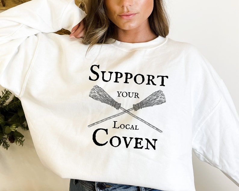 May include: White sweatshirt with black text that reads "Support Your Local Coven" with a graphic of two crossed broomsticks.