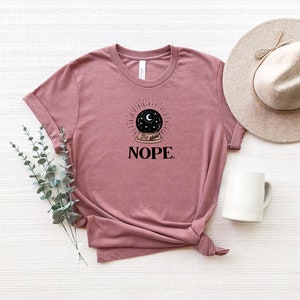 May include: A mauve t-shirt with a black and white illustration of a crystal ball with a crescent moon and stars inside. The text "NOPE." is printed below the illustration.