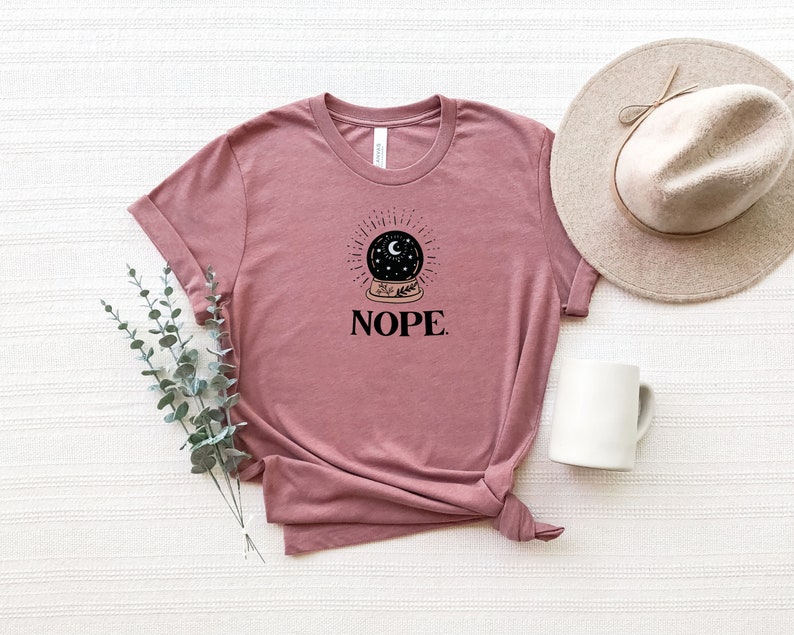 May include: A mauve t-shirt with a black and white illustration of a crystal ball with a crescent moon and stars inside. The text "NOPE." is printed below the illustration.
