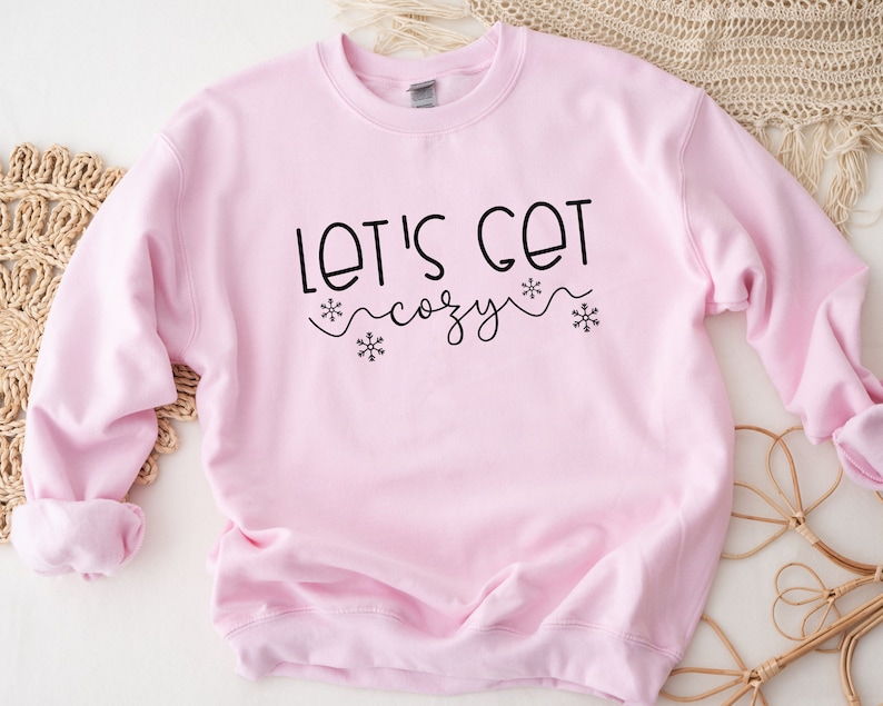 May include: A pink sweatshirt with the text "Let's Get Cozy" in black lettering with snowflake accents.