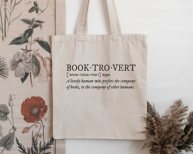 May include: A beige tote bag with a black print that reads "BOOK TRO VERT [BOOK-TRUH-VURT] noun. A lovely human who prefers the company of books, to the company of other humans."