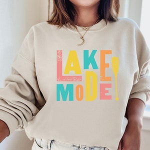 May include: A beige sweatshirt with the text "LAKE MODE" in colorful block letters and a paddle graphic. The letters are in shades of pink, yellow, blue, and orange.