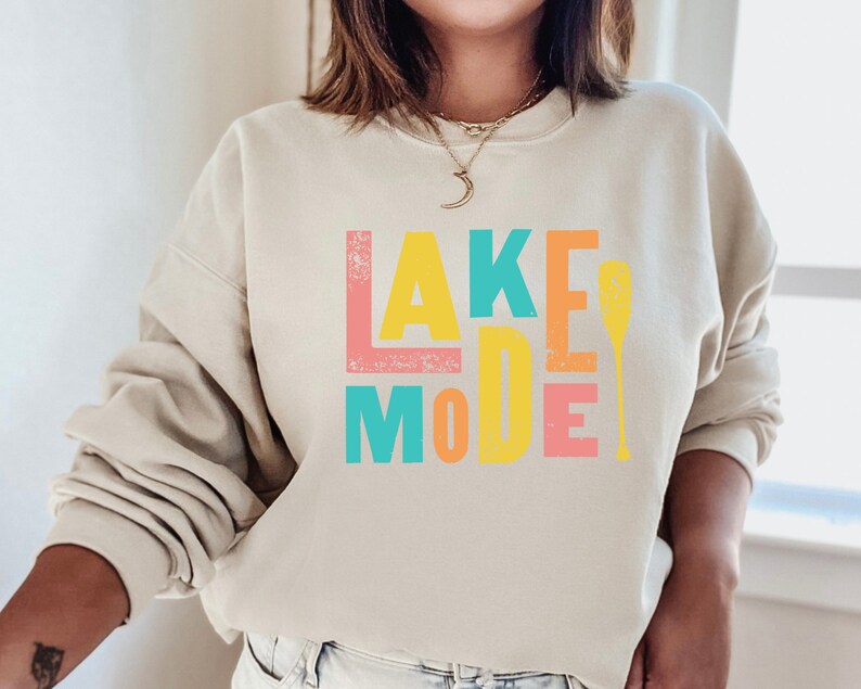 May include: A beige sweatshirt with the text "LAKE MODE" in colorful block letters and a paddle graphic. The letters are in shades of pink, yellow, blue, and orange.