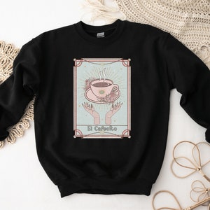 May include: Black sweatshirt with a graphic of a coffee cup with steam rising from it, surrounded by flowers and hands. The text "El Cafeito" is below the cup.