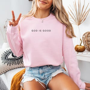 K&ouml;nnte beinhalten: Ein rosa Sweatshirt mit dem Schriftzug "GOD IS GOOD" in schwarzer Schrift.