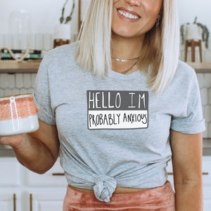 May include: A gray t-shirt with a white graphic that says "Hello I'm Probably Anxious". The shirt is tied at the waist and the person wearing it is holding a pink and white mug.
