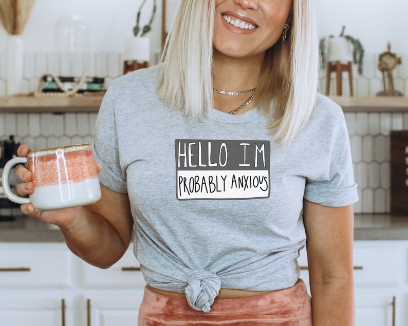 May include: A gray t-shirt with a white graphic that says "Hello I'm Probably Anxious". The shirt is tied at the waist and the person wearing it is holding a pink and white mug.