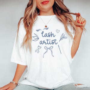 May include: White t-shirt with the text "lash artist" in blue lettering. The shirt also features a stylized illustration of eyelashes, a heart, a bow, scissors, and a lash applicator.