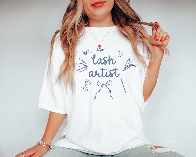 May include: White t-shirt with the text "lash artist" in blue lettering. The shirt also features a stylized illustration of eyelashes, a heart, a bow, scissors, and a lash applicator.