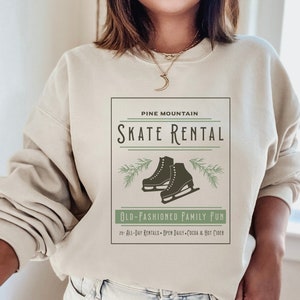 K&ouml;nnte beinhalten: Ein beiger Pullover mit einem grafischen Design, das den Text "Pine Mountain Skate Rental" und ein Paar Schlittschuhe zeigt. Unter dem Grafik befindet sich der Text "Old-Fashioned Family Fun" sowie der Text "25+ All-Day Rentals - Open Daily - Cocoa & Hot Cider".