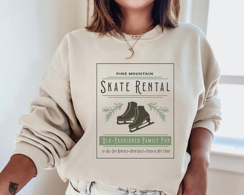 K&ouml;nnte beinhalten: Ein beiger Pullover mit einem grafischen Design, das den Text "Pine Mountain Skate Rental" und ein Paar Schlittschuhe zeigt. Unter dem Grafik befindet sich der Text "Old-Fashioned Family Fun" sowie der Text "25+ All-Day Rentals - Open Daily - Cocoa & Hot Cider".