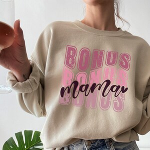 May include: A beige sweatshirt with a pink and white graphic design that says "Bonus mama" in a retro style.