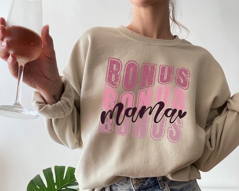 May include: A beige sweatshirt with a pink and white graphic design that says "Bonus mama" in a retro style.