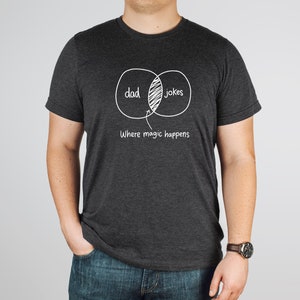 May include: A dark gray t-shirt with a white graphic of a Venn diagram. The diagram shows the overlap between "dad" and "jokes" with the text "Where magic happens" below.
