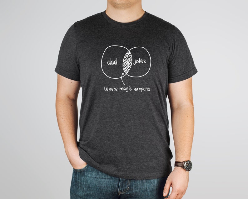 May include: A dark gray t-shirt with a white graphic of a Venn diagram. The diagram shows the overlap between "dad" and "jokes" with the text "Where magic happens" below.