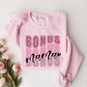 May include: Pink sweatshirt with the text "BONUS mama BONUS" in a retro font with a distressed effect.