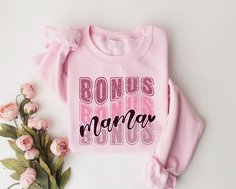 May include: Pink sweatshirt with the text "BONUS mama BONUS" in a retro font with a distressed effect.
