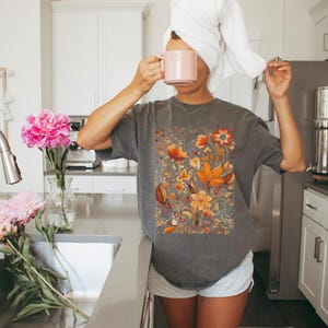 May include: Gray t-shirt with a floral design in shades of orange, yellow, and red. The image also shows a pink mug, a white towel, and pink flowers in a vase. The word "Pepper" is visible.