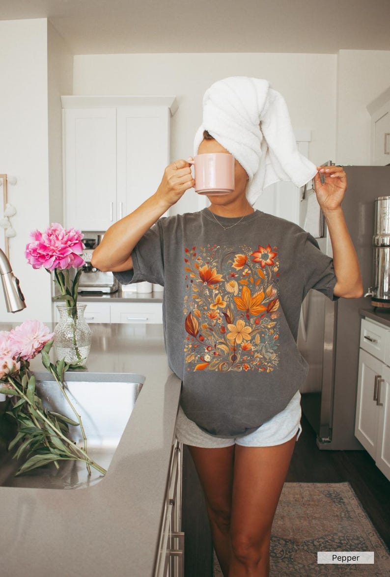 May include: Gray t-shirt with a floral design in shades of orange, yellow, and red. The image also shows a pink mug, a white towel, and pink flowers in a vase. The word "Pepper" is visible.
