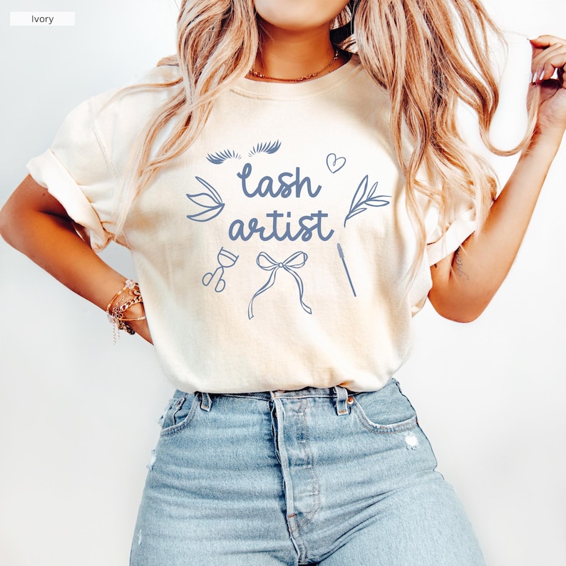 May include: A white t-shirt with the text "lash artist" in blue lettering. The shirt also features a blue illustration of eyelashes, a heart, a bow, and other lash-related tools.
