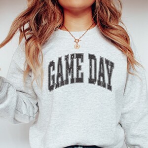 May include: A gray sweatshirt with the text "GAME DAY" printed in black, with a distressed vintage look. The sweatshirt is worn by a person with long blonde hair.