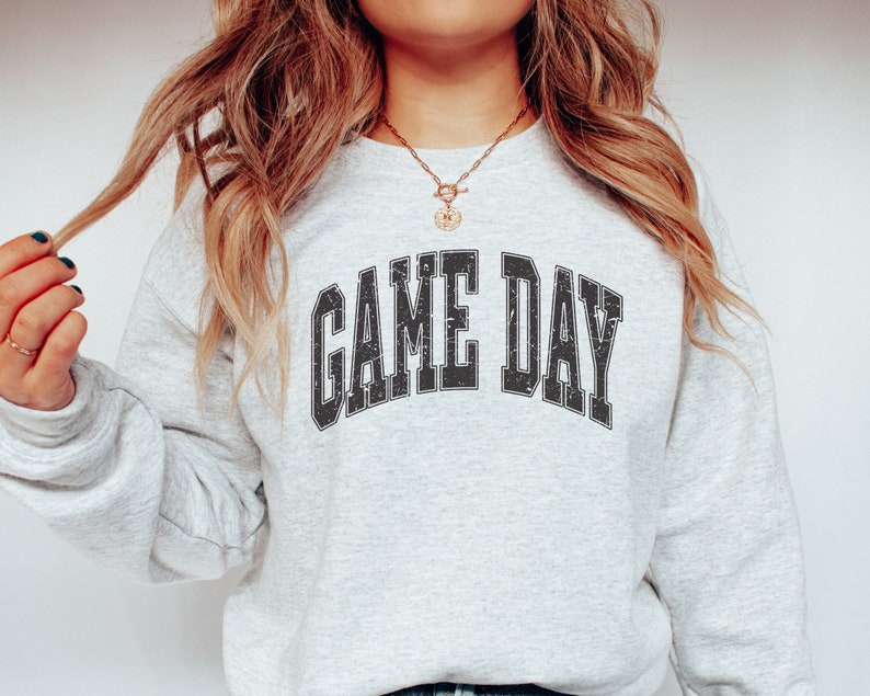 May include: A gray sweatshirt with the text "GAME DAY" printed in black, with a distressed vintage look. The sweatshirt is worn by a person with long blonde hair.