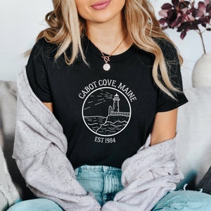 Puede incluir: Una camiseta negra con un gr&aacute;fico blanco de un faro y el texto "Cabot Cove, Maine Est 1984".