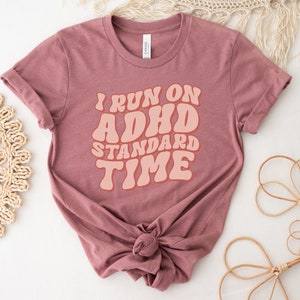 May include: A mauve t-shirt with the text "I run on ADHD standard time" in a retro font style. The text is in a peach colour.