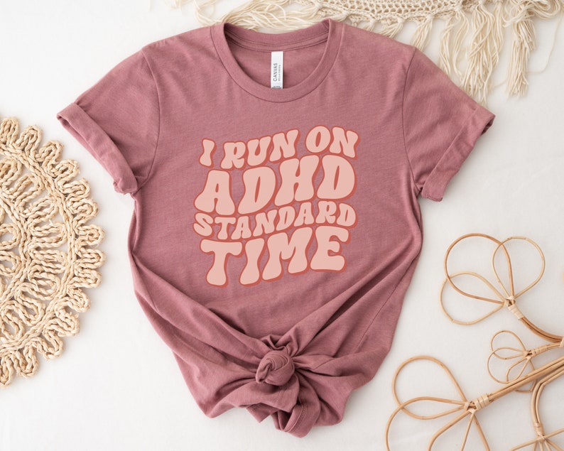 May include: A mauve t-shirt with the text "I run on ADHD standard time" in a retro font style. The text is in a peach colour.