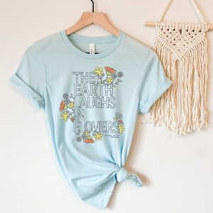 Pode incluir: Camiseta azul claro com um design gr&aacute;fico de flores e o texto "The Earth Laughs in Flowers".