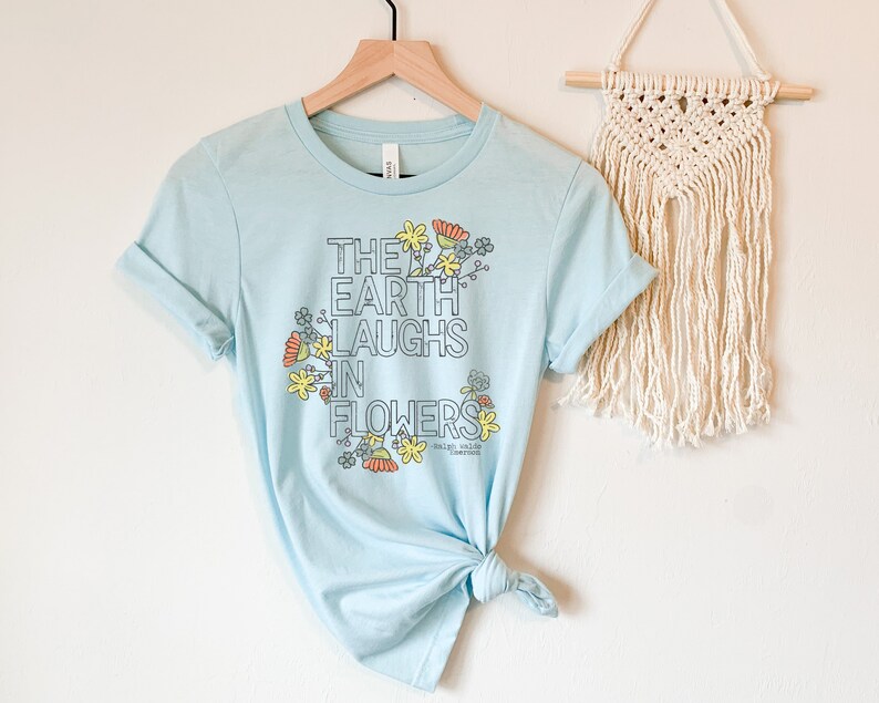 Pode incluir: Camiseta azul claro com um design gr&aacute;fico de flores e o texto "The Earth Laughs in Flowers".