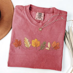 May include: A maroon t-shirt with a line of colorful fall leaves printed on the front. The shirt has a tag that reads "Comfort Colors" and "S - P - CH".