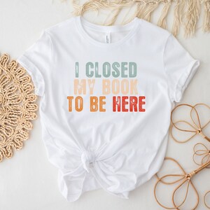 Puede incluir: Camiseta blanca con un gr&aacute;fico de estilo retro que dice "Cerr&eacute; mi libro para estar aqu&iacute;".