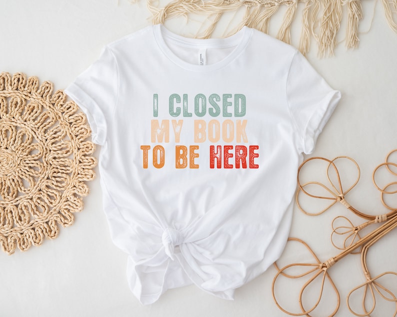 Puede incluir: Camiseta blanca con un gr&aacute;fico de estilo retro que dice "Cerr&eacute; mi libro para estar aqu&iacute;".