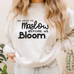 May include: A white sweatshirt with the text "We have to Maslow before we Bloom." printed in black.