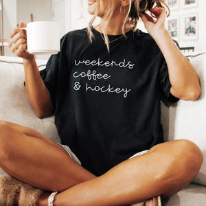 May include: Black t-shirt with the words "weekends coffee & hockey" in white script. The shirt is worn with light-colored shorts. A white mug is held in the hand. The person is sitting on a light-colored couch.