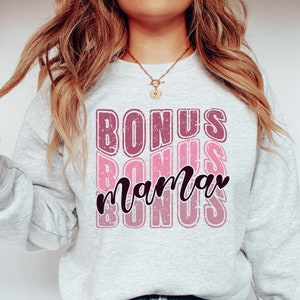 May include: A gray sweatshirt with the text "BONUS" repeated three times in a pink and purple gradient. The word "mama" is written in black cursive between the words "BONUS".
