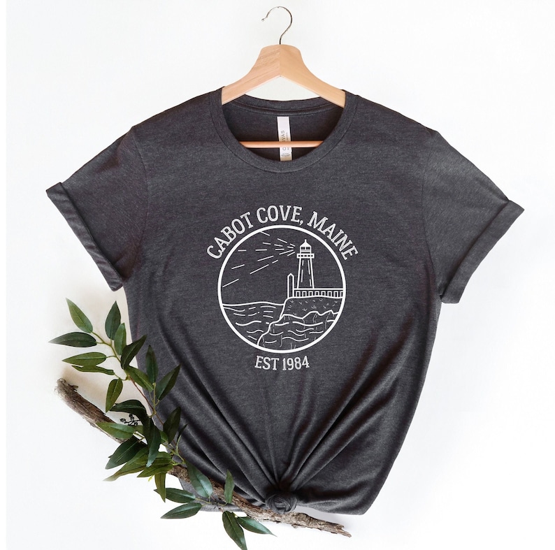 Puede incluir: Camiseta gris oscuro con un gr&aacute;fico blanco de un faro en un c&iacute;rculo con el texto "Cabot Cove, Maine Est 1984".