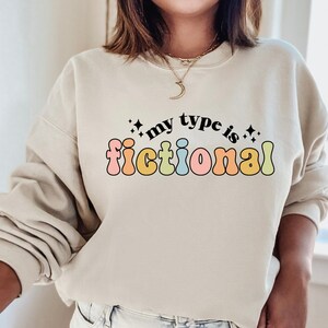 May include: A light beige sweatshirt with the text "my type is fictional" in rainbow-colored letters. The text is in a retro font style.